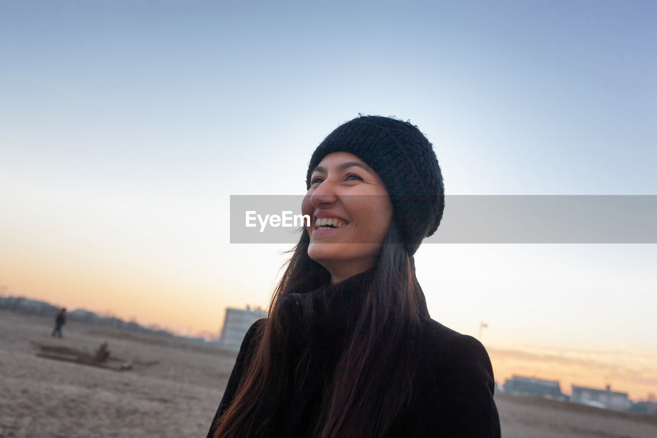 PORTRAIT OF SMILING YOUNG WOMAN AT BEACH AGAINST SKY