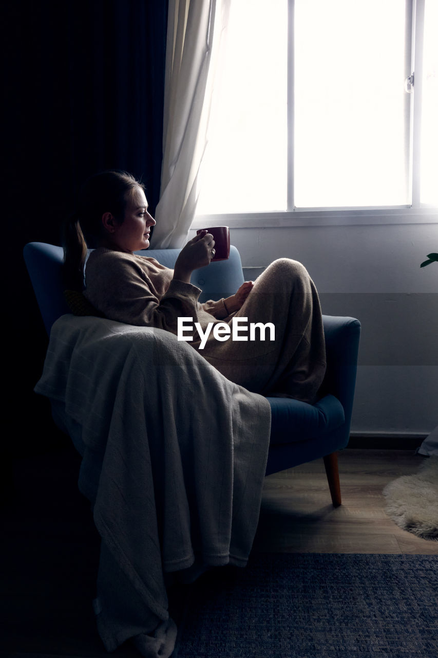 Young woman sitting on sofa at home and drinking coffee or tea