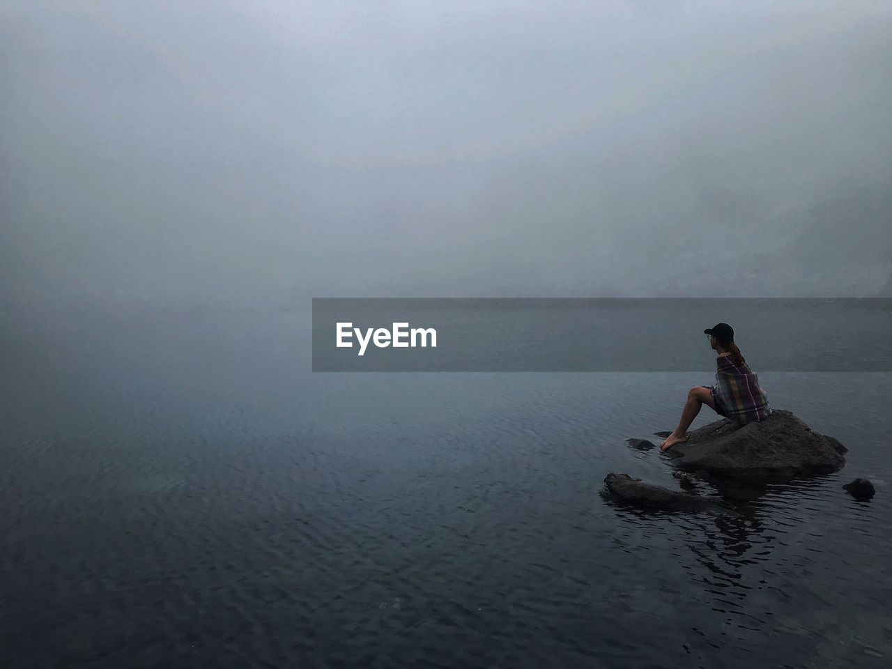 Side view of woman sitting on rock in lake during foggy weather