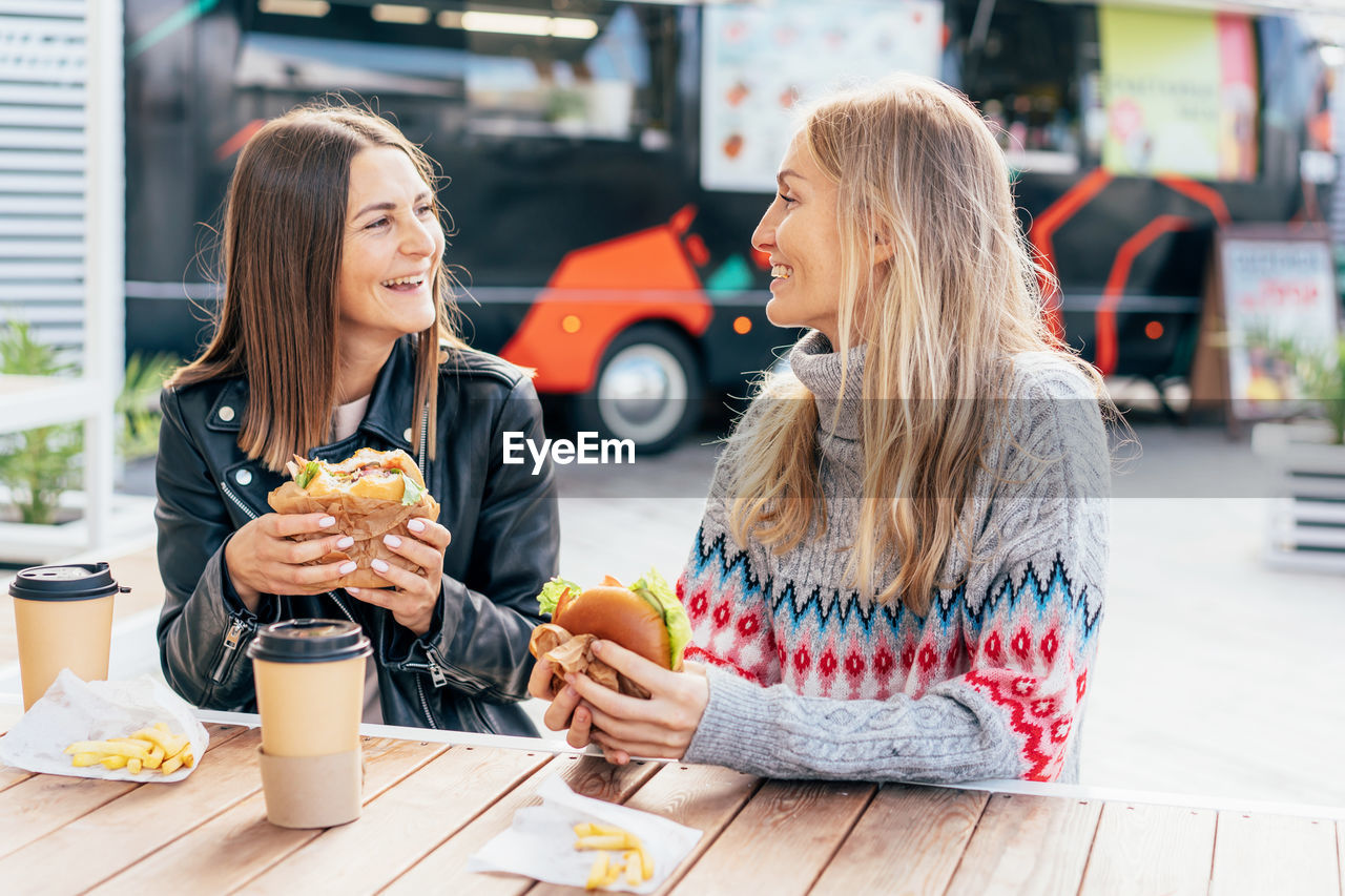 Two middle-aged women eat fast food and chat at a street food market.