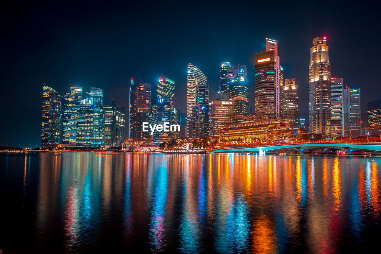 Illuminated buildings by river against sky at night