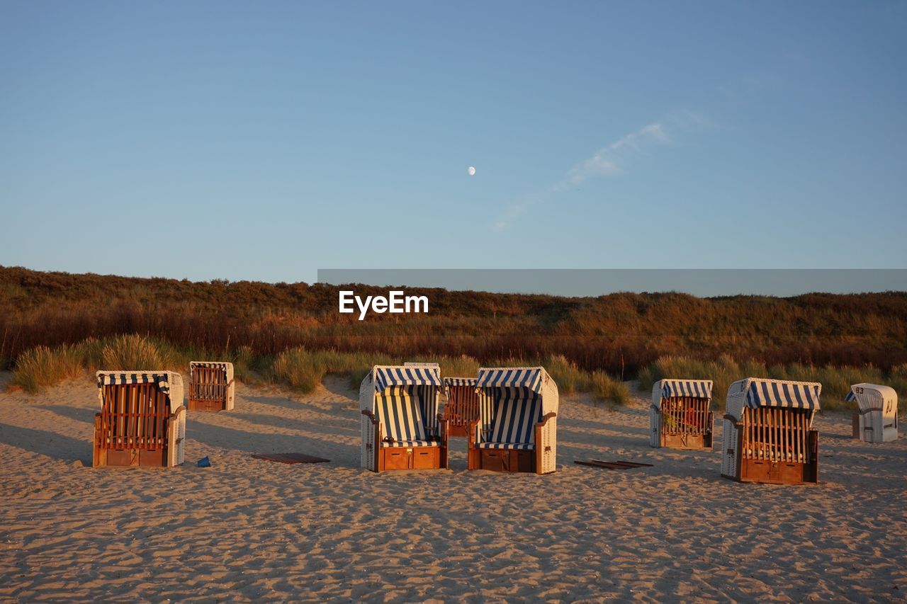 Hooded beach chairs on sand against blue sky during sunset