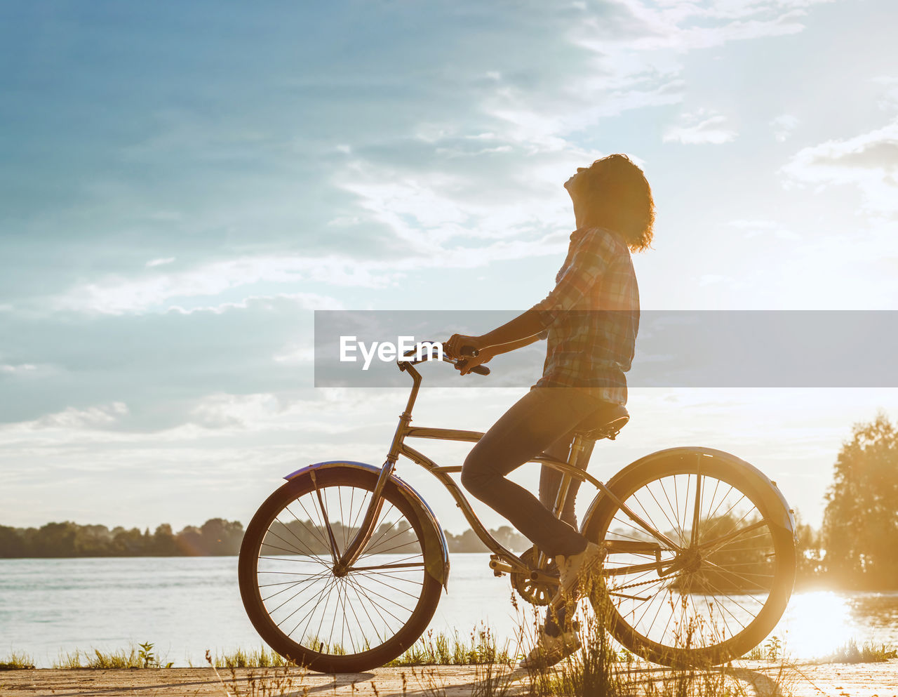 Side view of woman riding bicycle by lake against sky at sunset