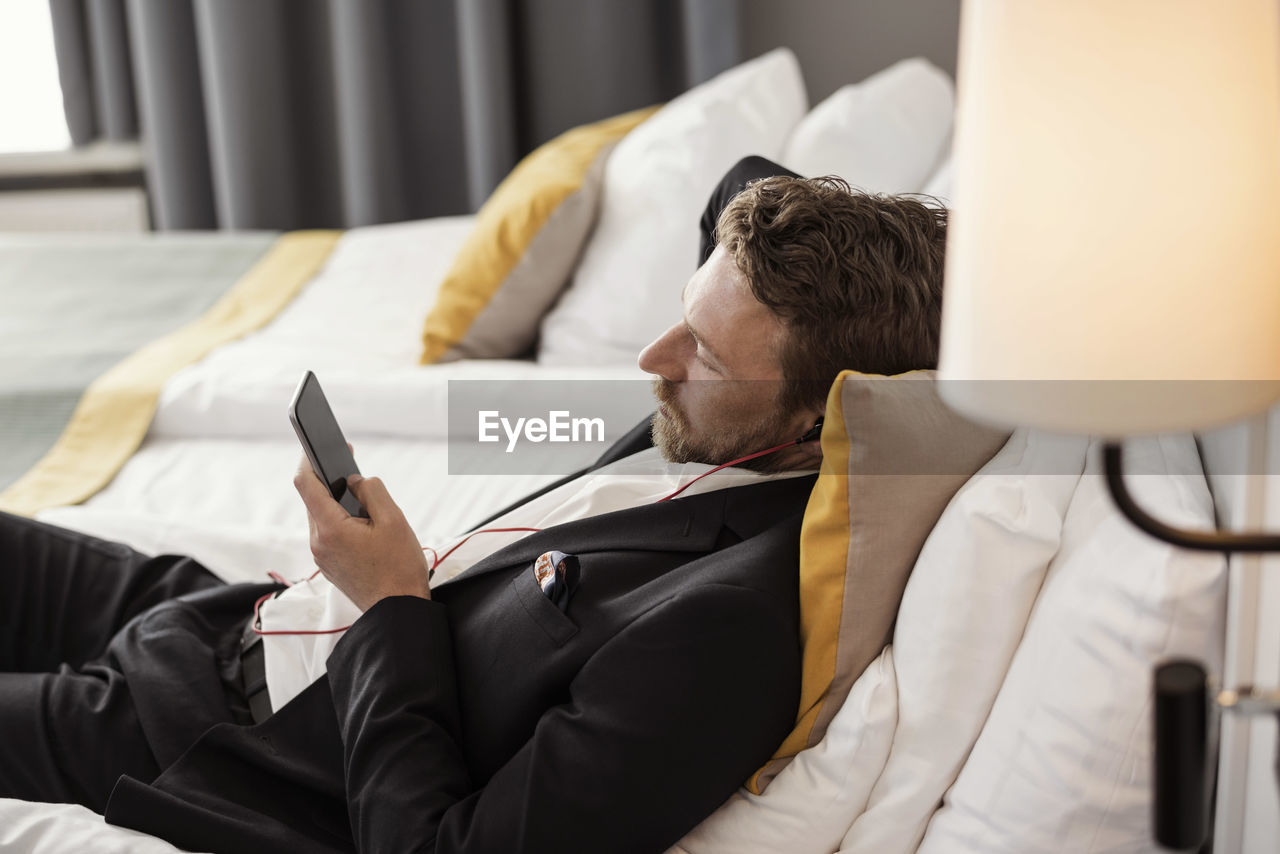 High angle view of businessman using mobile phone listening to music while lying on bed at hotel room