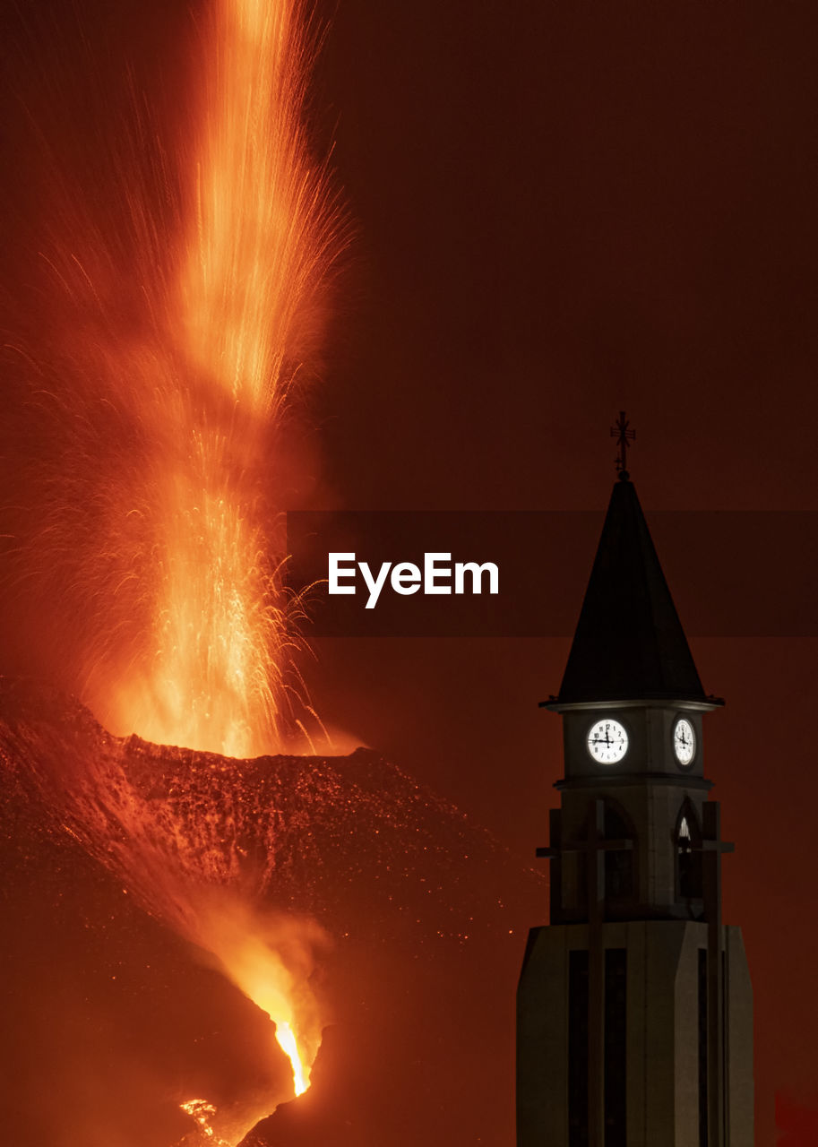 Dramatic scenery of church clock tower silhouette on mountain slope against powerful eruption of cumbre vieja volcano at night in la palma island