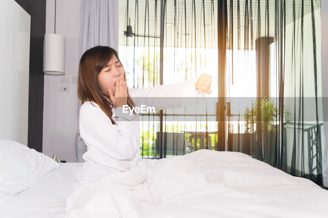 Young woman yawning while sitting on bed at home