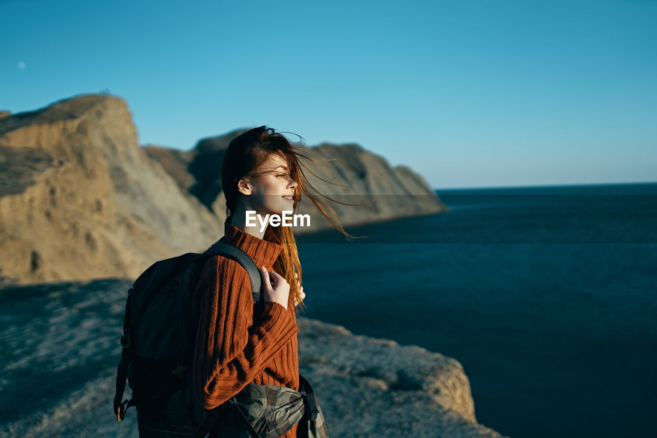 YOUNG WOMAN STANDING ON ROCK AT SEA SHORE AGAINST SKY