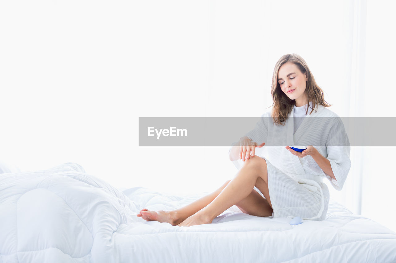 Young woman applying moisturizer while sitting on bed