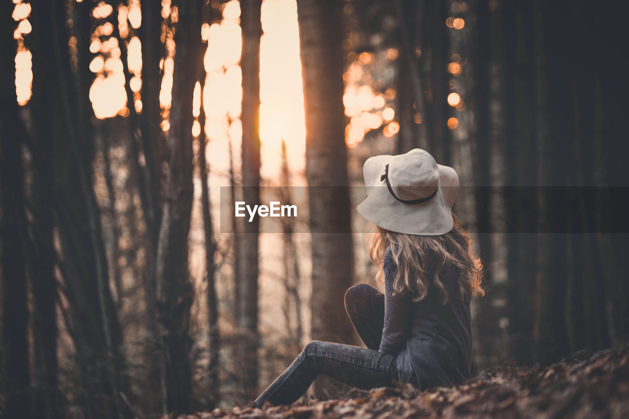 Woman in hat sitting on field against trees during sunset