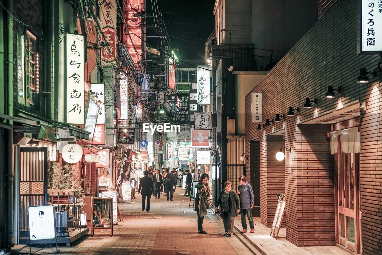 PEOPLE WALKING ON ILLUMINATED STREET AMIDST BUILDINGS AT NIGHT