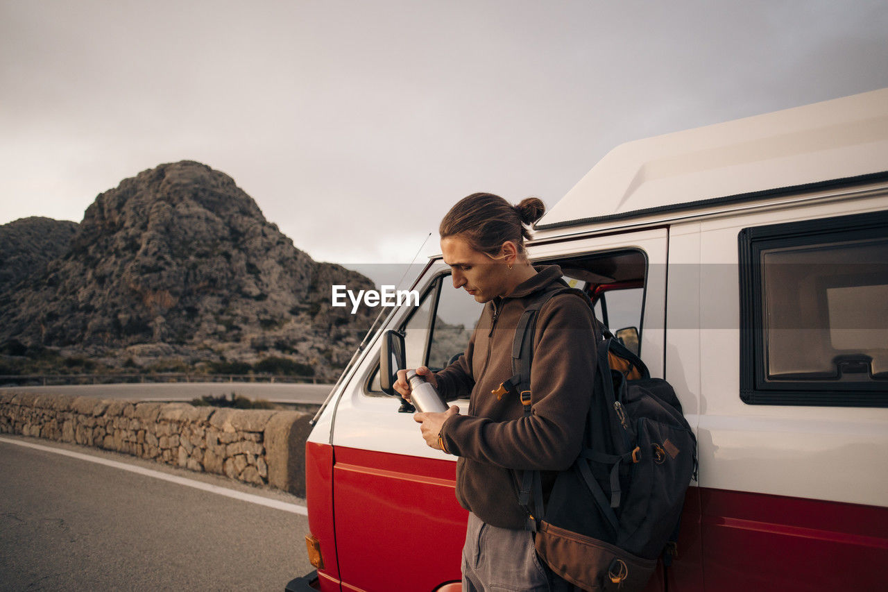 Man with backpack opening water bottle while standing near van on vacation