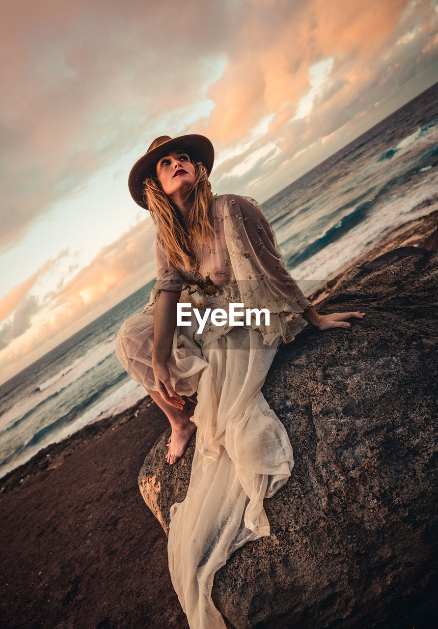 Woman wearing hat while sitting on rock at beach during sunset