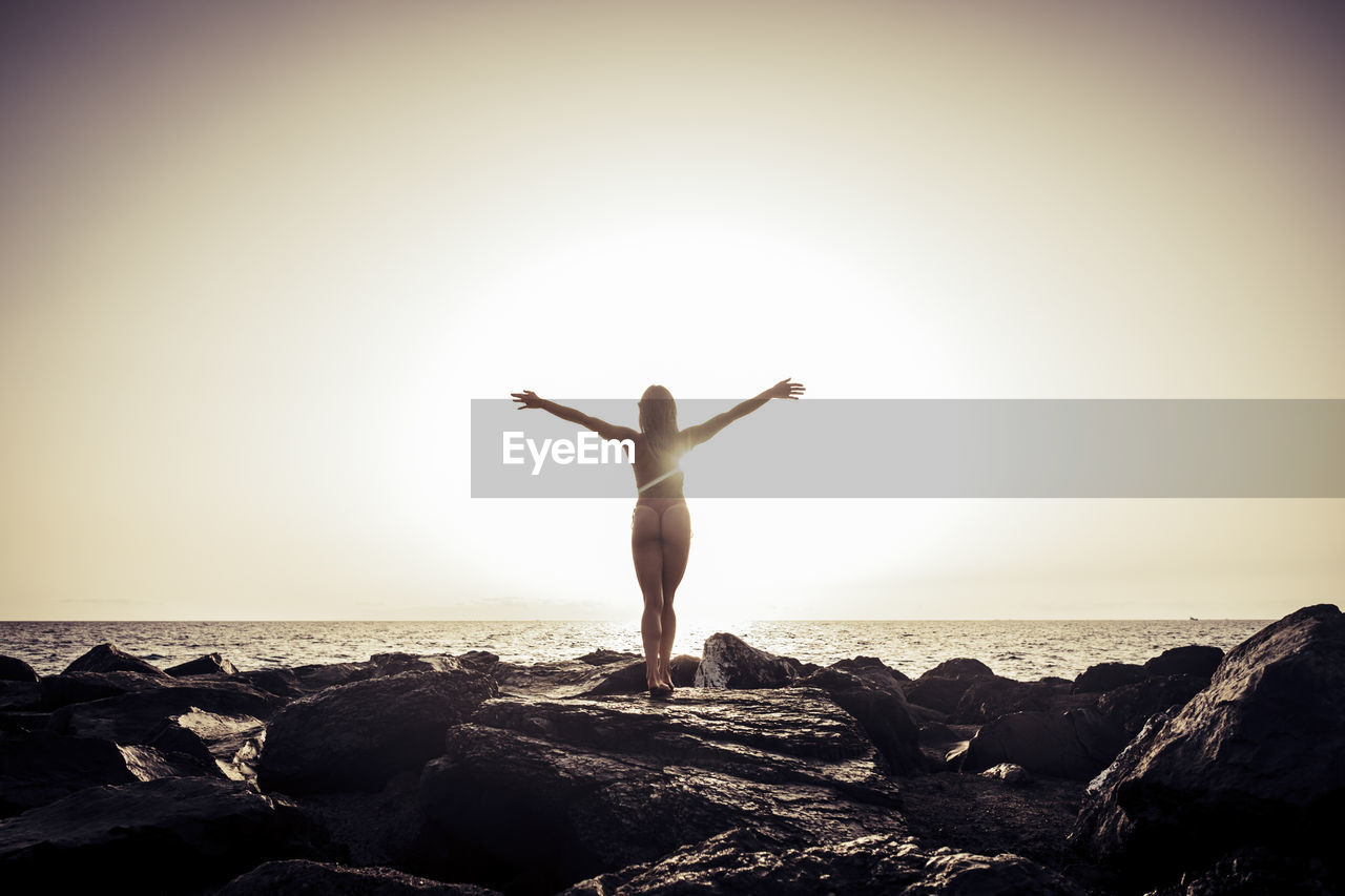 Rear view of woman with arms outstretched on rocky shore against clear sky