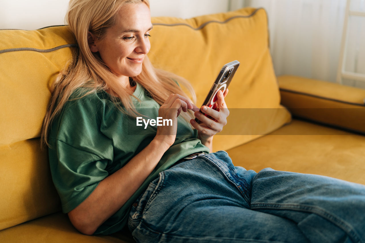 Caucasian middle aged woman browsing media in mobile phone while sitting on yellow sofa at home.