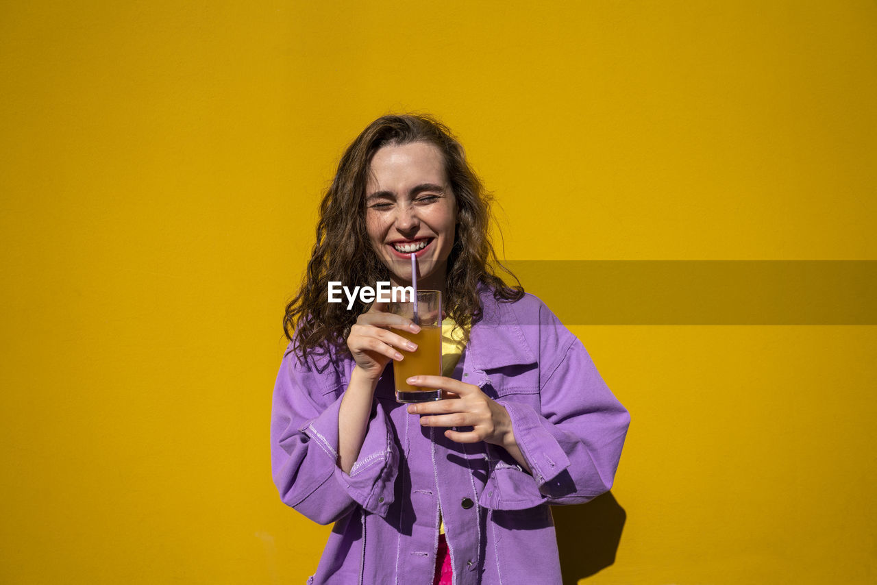 Cheerful woman with eyes closed drinking juice in front of yellow wall