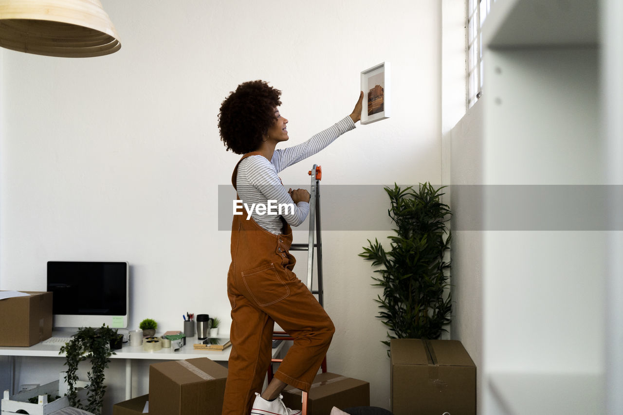 Afro woman putting picture frame on wall while climbing on ladder in new apartment