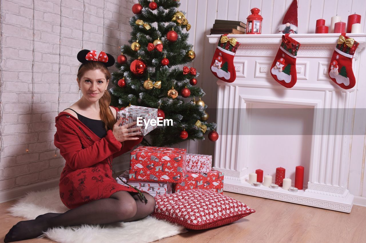 Portrait of young woman holding gift box by christmas tree at home