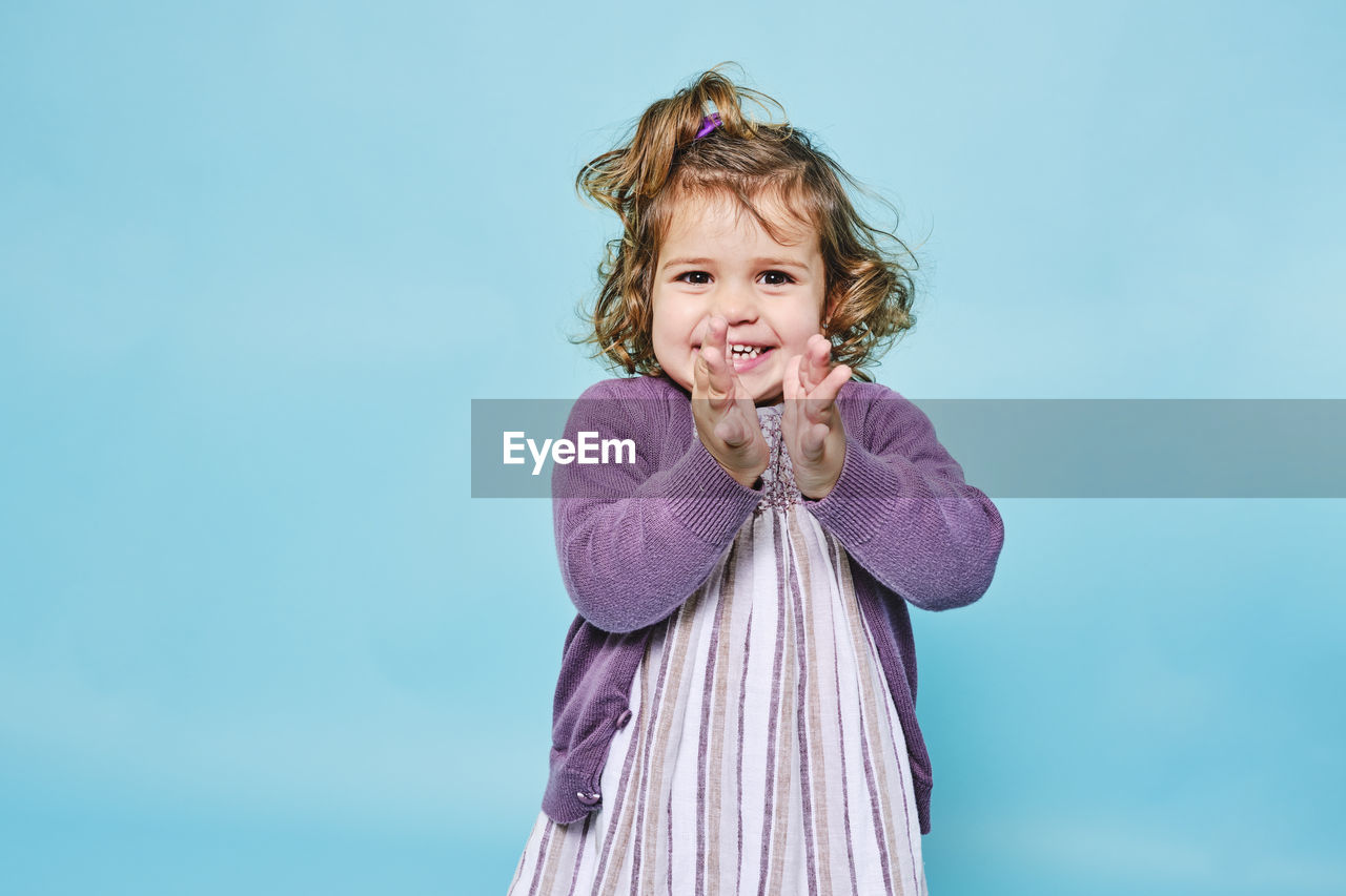 Cheerful small child in purple dress and knitted cardigan smiling at camera and clapping while standing alone against light blue background in modern studio