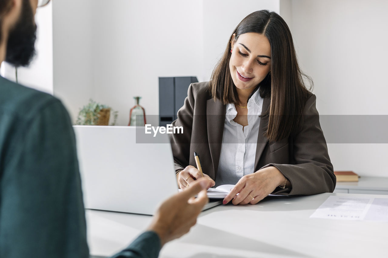 Smiling recruiter interviewing candidate and writing in diary at desk