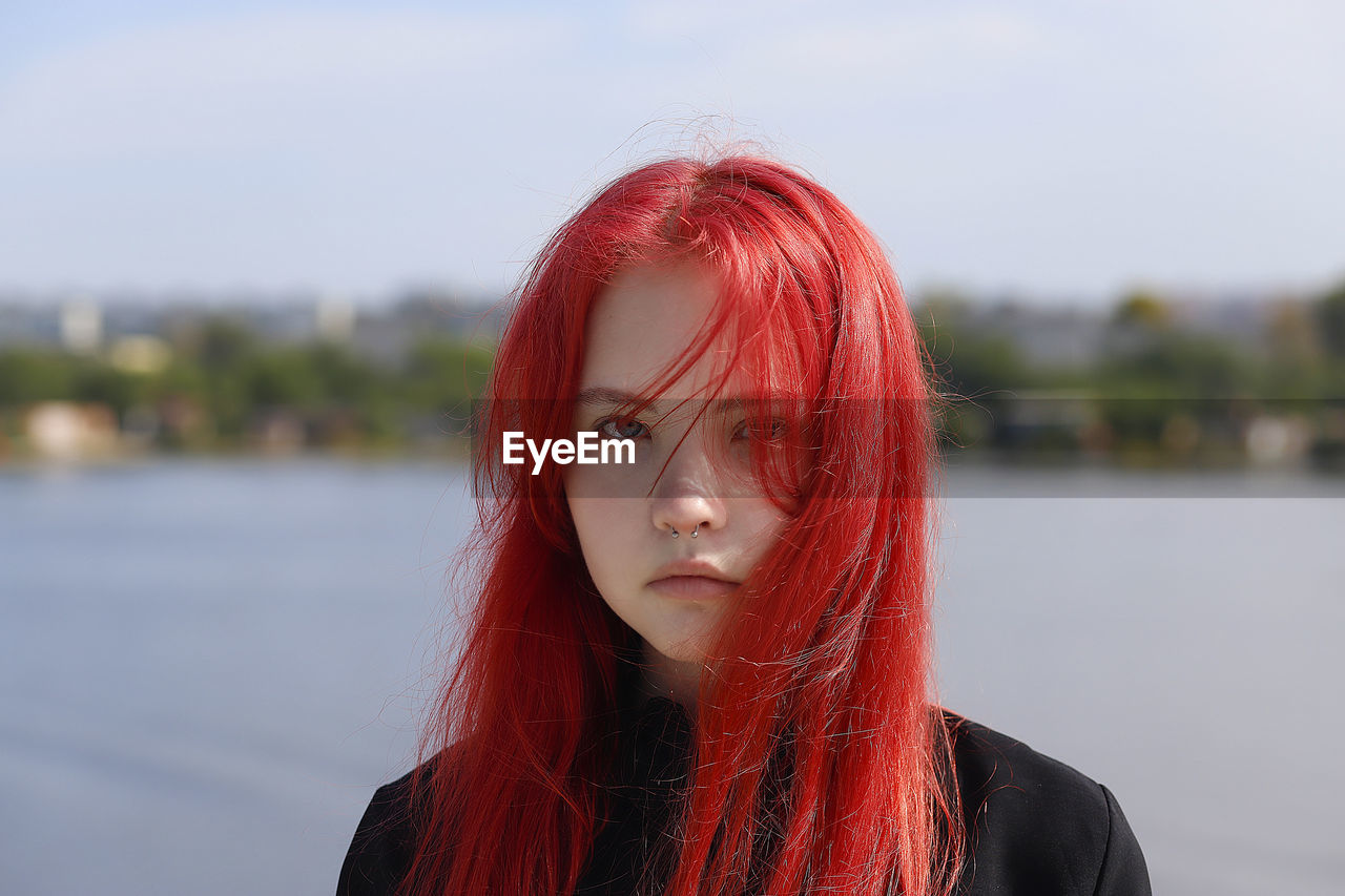 A young girl with a pierced nose and red hair stands on  the background of a river on a sunny day