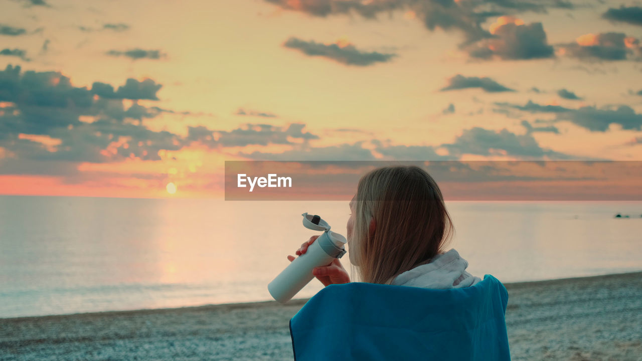 Woman drinking water against sky during sunset