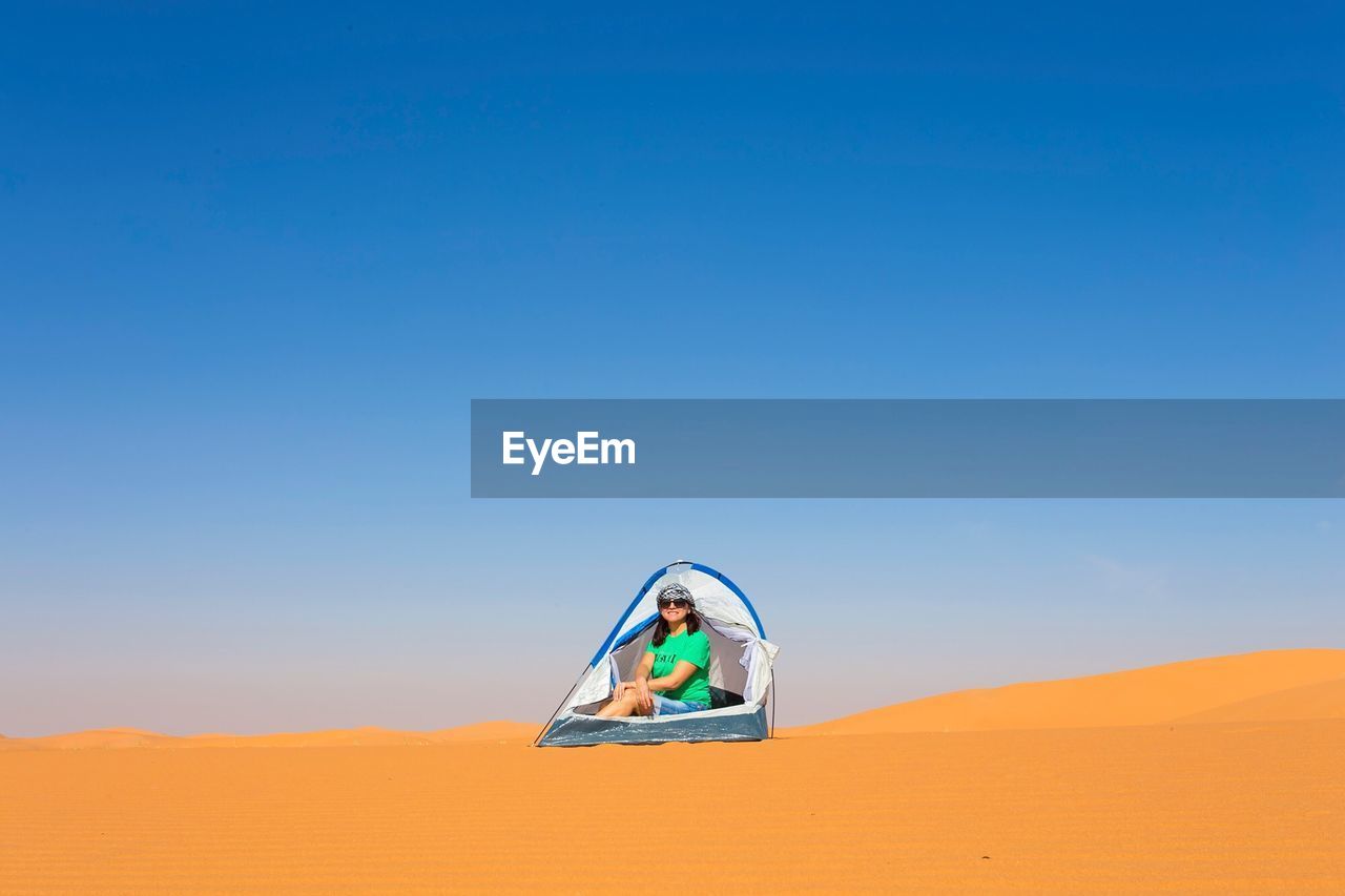 Mature woman sitting in tent on sand at desert against clear blue sky