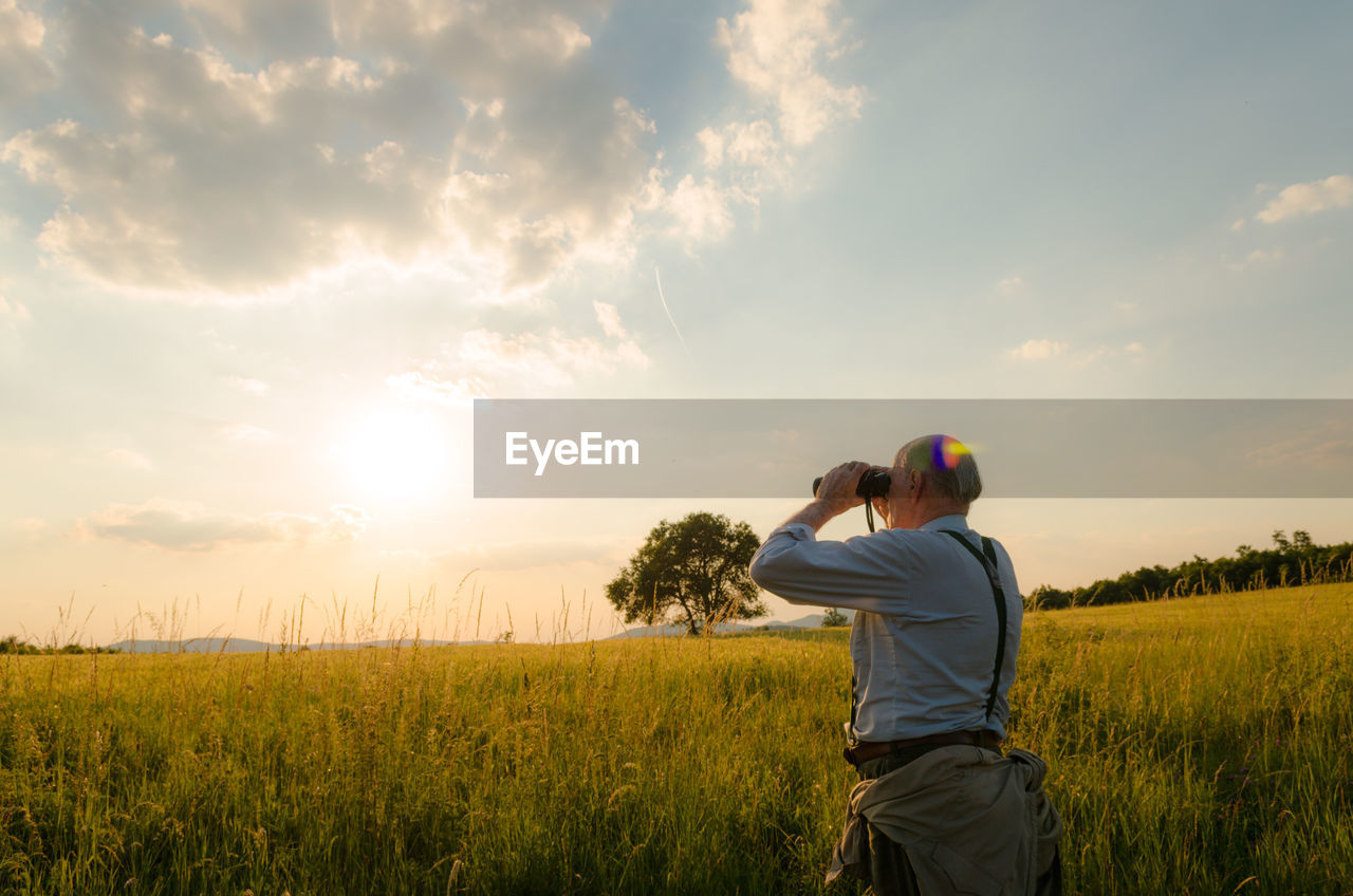 Rear view of man using binoculars on field against sky during sunset