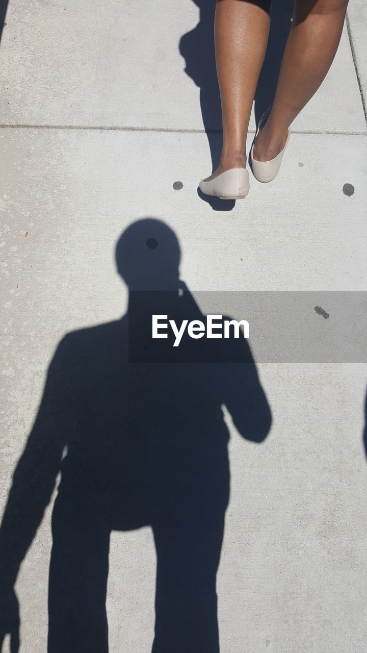 Low section of woman with man shadow on footpath during sunny day