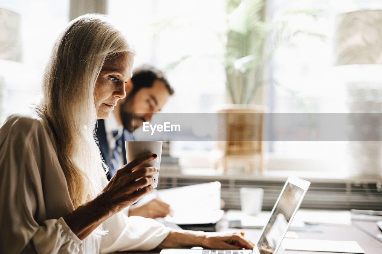 Mature businesswoman holding coffee mug using laptop by businessman at desk in office