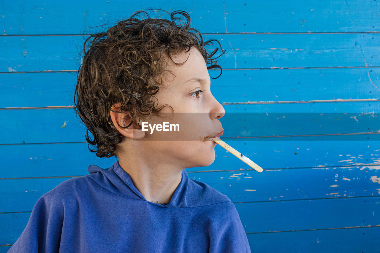 Close-up of boy eating popsicle looking away against blue wall