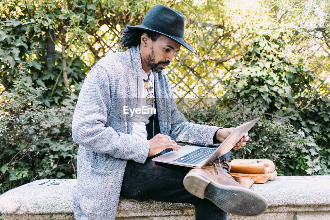 Adult hispanic man in trendy clothes and hat browsing netbook while sitting on stone bench and working remotely from park