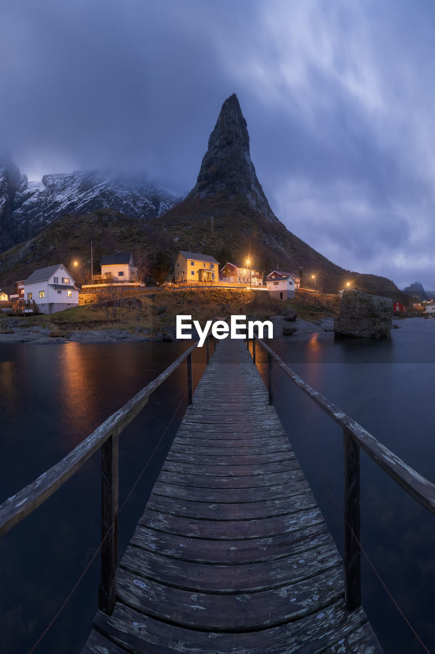 Amazing view of wooden footbridge crossing calm river and leading to illuminated village with cottages located at bottom of highlands in reine , lofoten islands in norway in evening