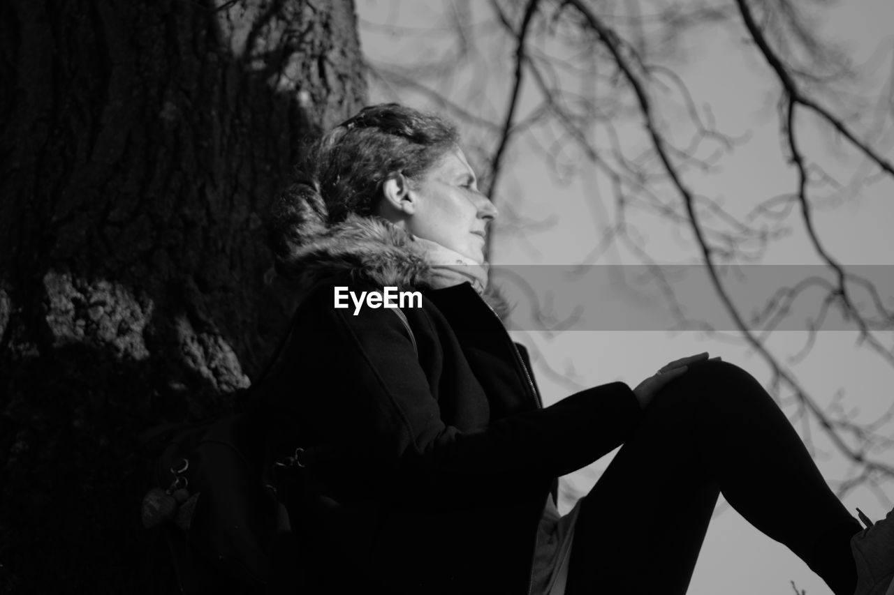 Side view of woman looking away while sitting against tree trunk