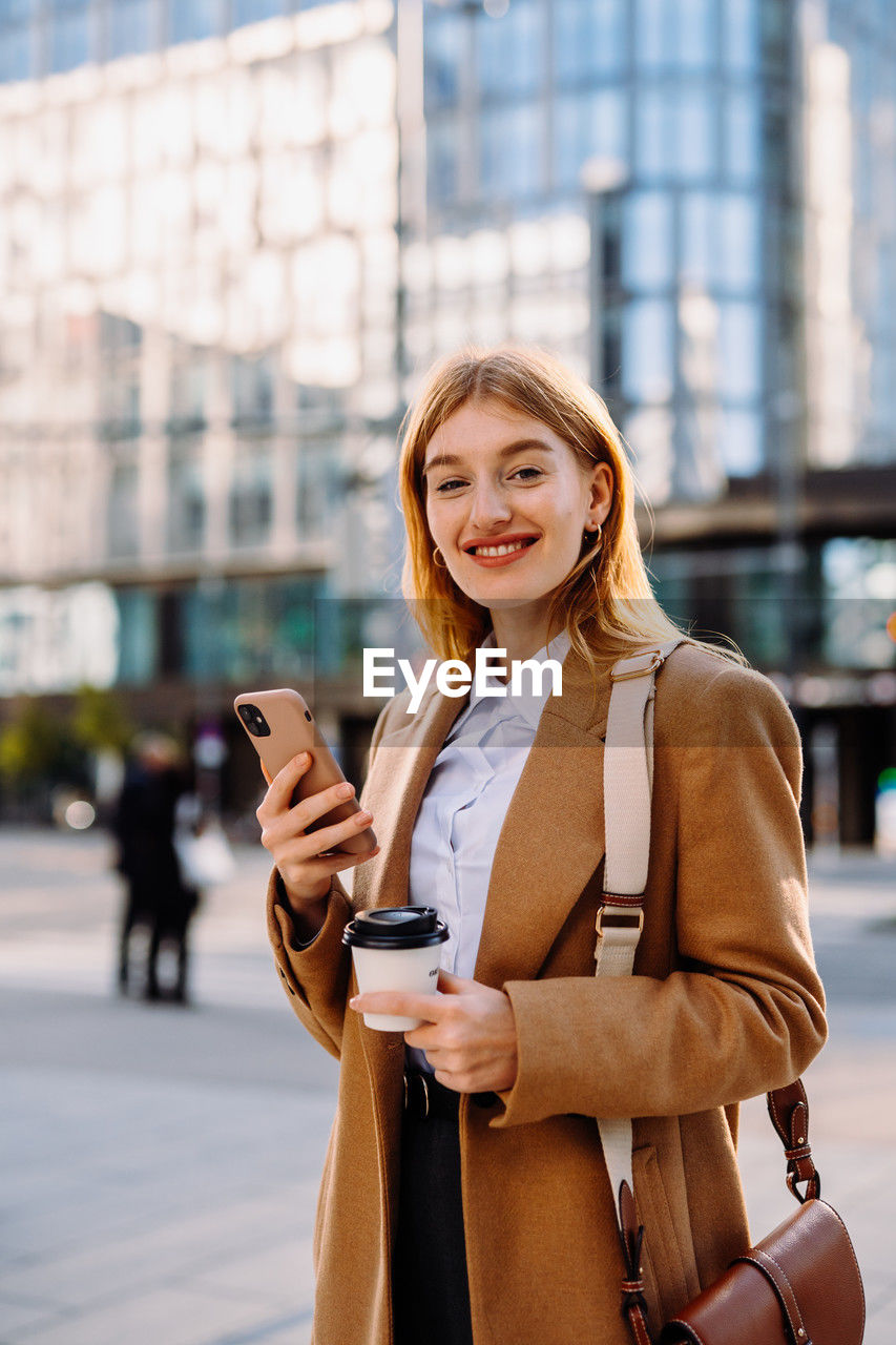 The woman in a blazer is smiling, holding a cup of coffee and a cell phone