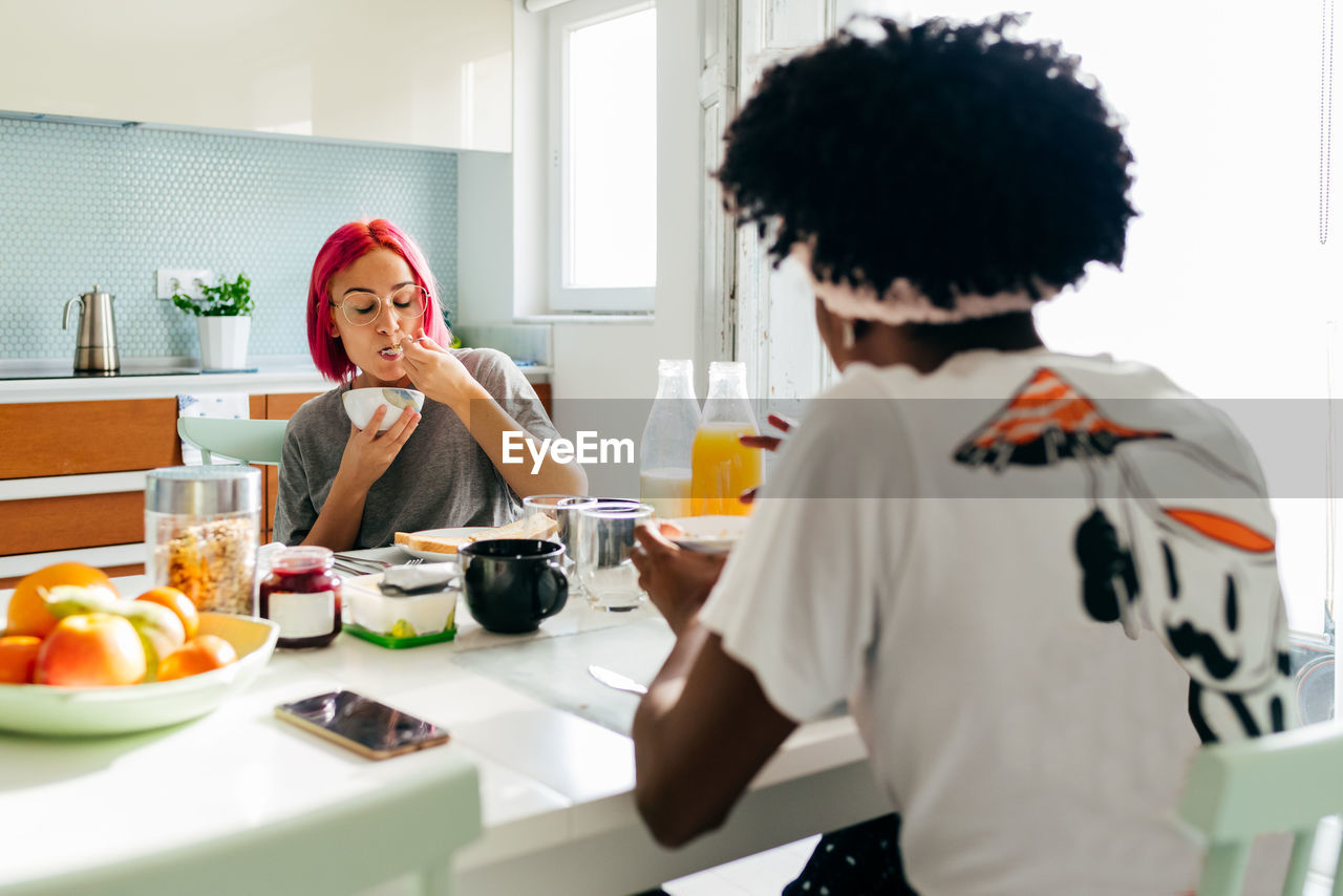 Relaxed young woman with dyed hair sitting near table and eating yummy dish near african american friend in cozy apartment