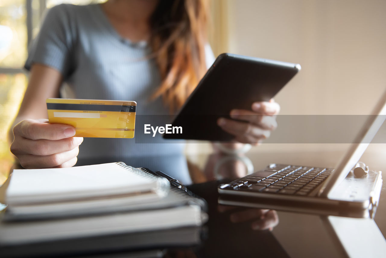 Female use credit cards to conduct financial transactions through tablet and laptop at home office.