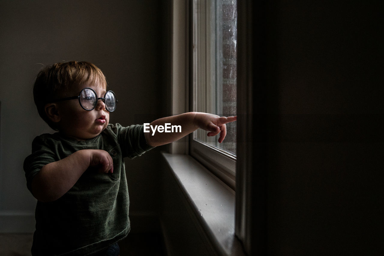Toddler boy wearing round glasses standing by window pointing outside