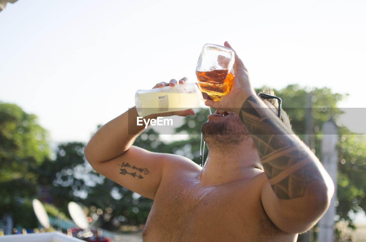 Midsection of shirtless man drinking glass against sky