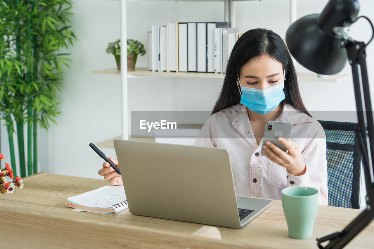 Businesswoman wearing mask using laptop and phone while sitting on table at office