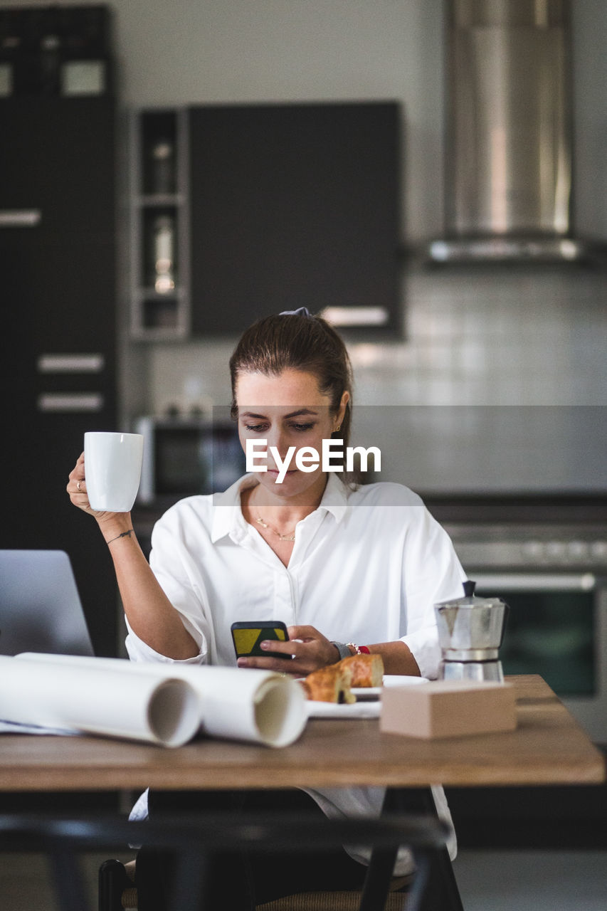 Female freelancer using phone while sitting with coffee cup at home