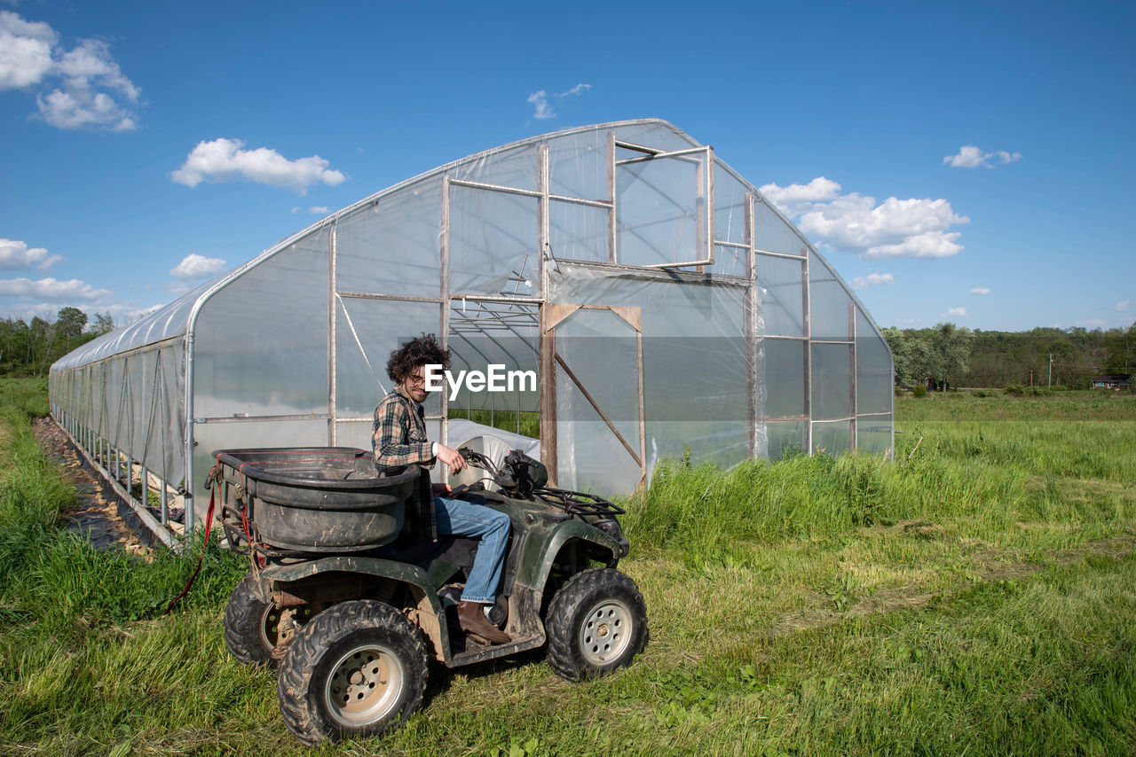 Young farmer on off road vehicle in sunny field by vegetable greenhouse