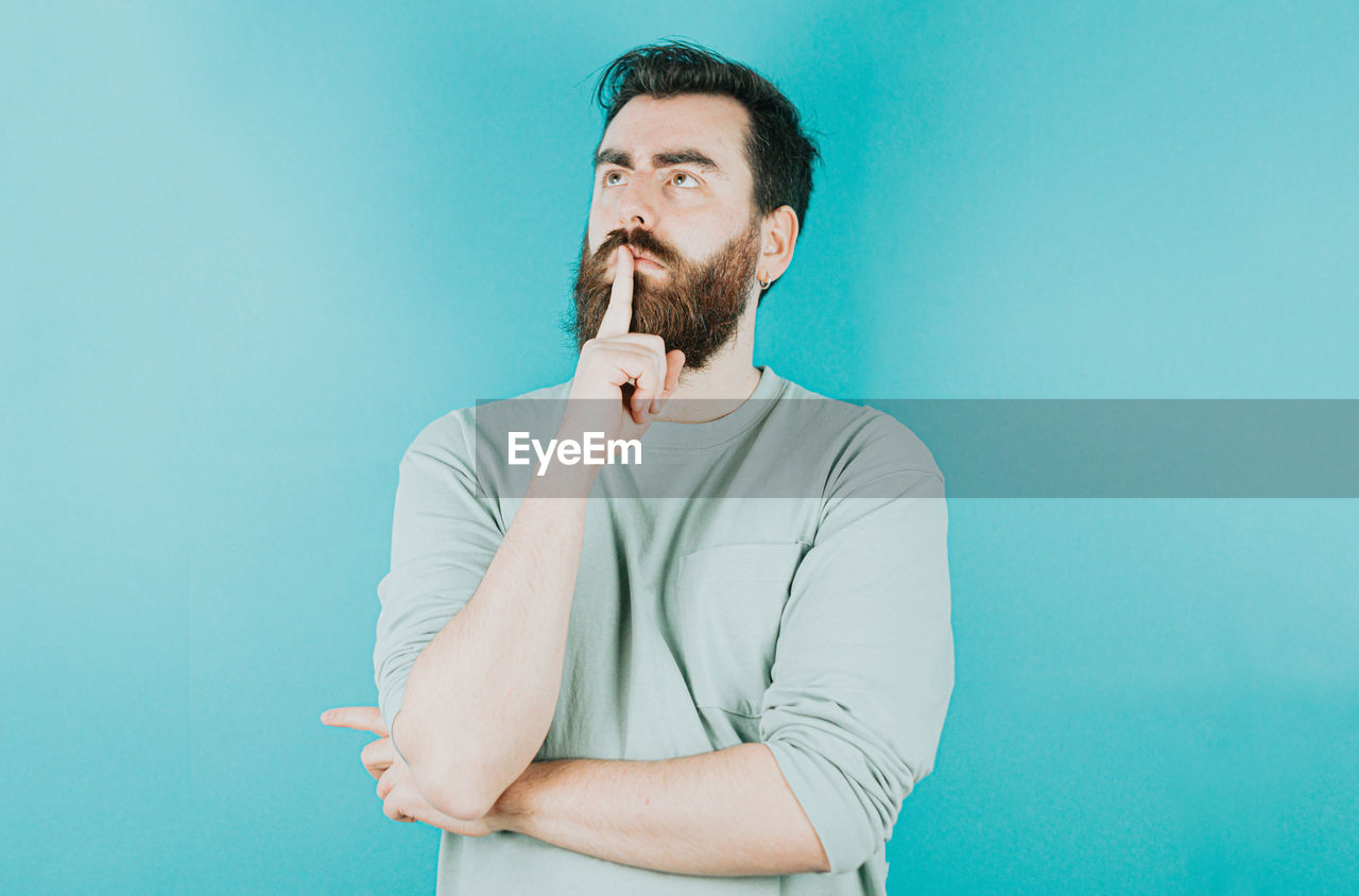 one person, adult, beard, facial hair, colored background, men, blue, indoors, waist up, studio shot, portrait, blue background, young adult, person, standing, looking, copy space, casual clothing, contemplation, businessman, looking away, emotion, button down shirt, clothing, business, lifestyles, front view, brown hair, wall - building feature, photo shoot