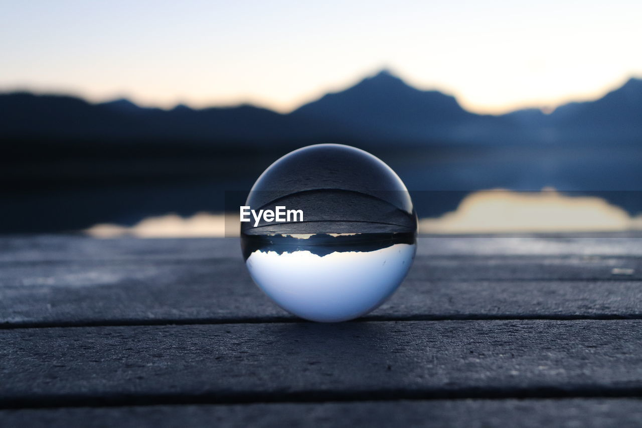 blue, reflection, water, nature, sphere, sky, no people, crystal ball, black, focus on foreground, scenics - nature, white, sea, outdoors, mountain, single object, environment, close-up, light, sunset, beauty in nature, tranquility, land, sunlight, selective focus, shape, day, landscape, wood, tranquil scene, transparent, beach, glass, travel
