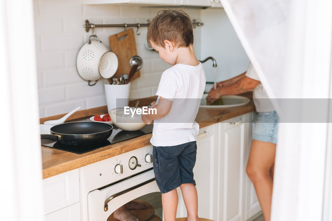 Cute toddler boy with mother cooking breakfast with puncakes together in bright kitchen at home