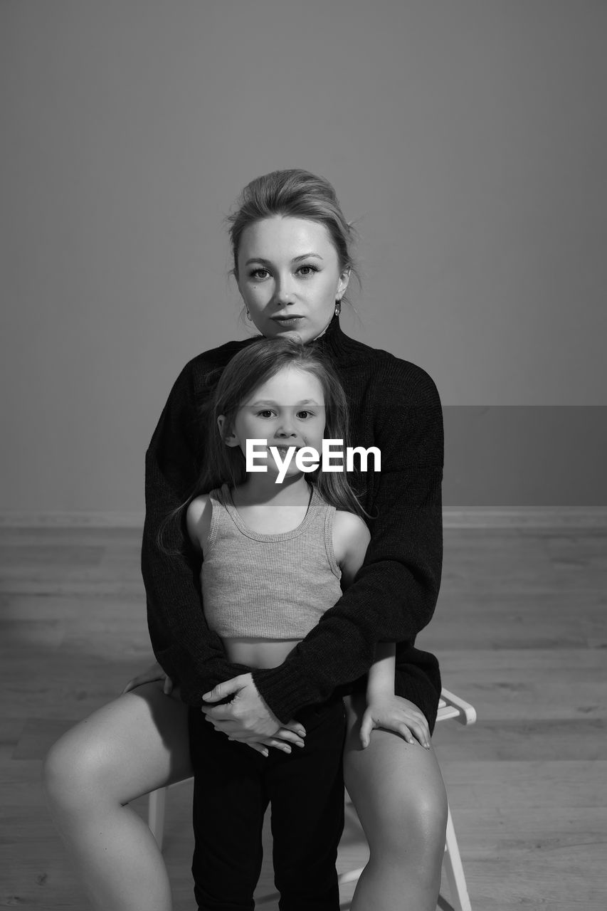 Mother in a black sweater hugs her five-year-old daughter sitting on a chair in the room in front