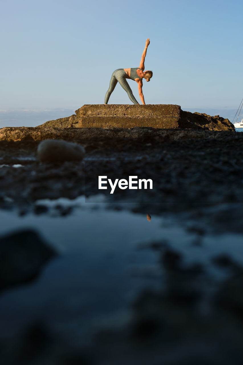 Side view of slim female in sportswear doing revolved triangle asana while practicing yoga on rocky seashore