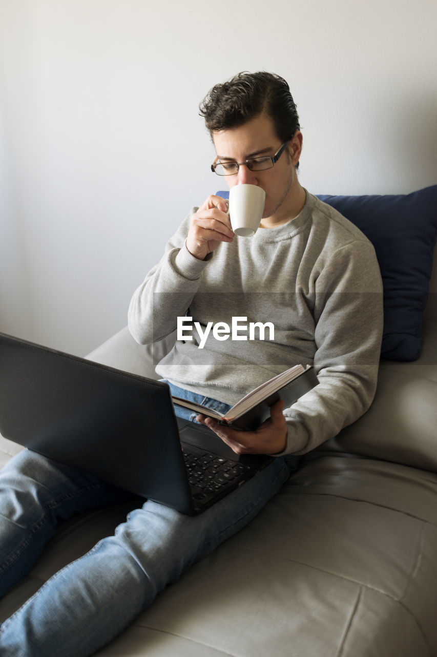 Young man sitting on bed using laptop and looking at a book