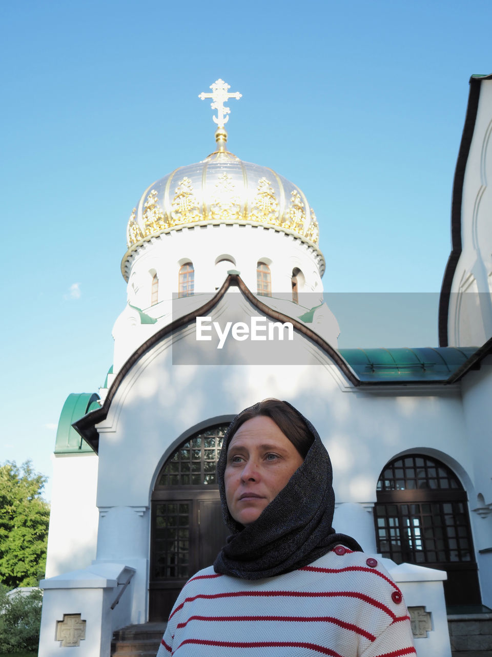 A girl of european appearance is standing in a kerchief at a white orthodox church. 