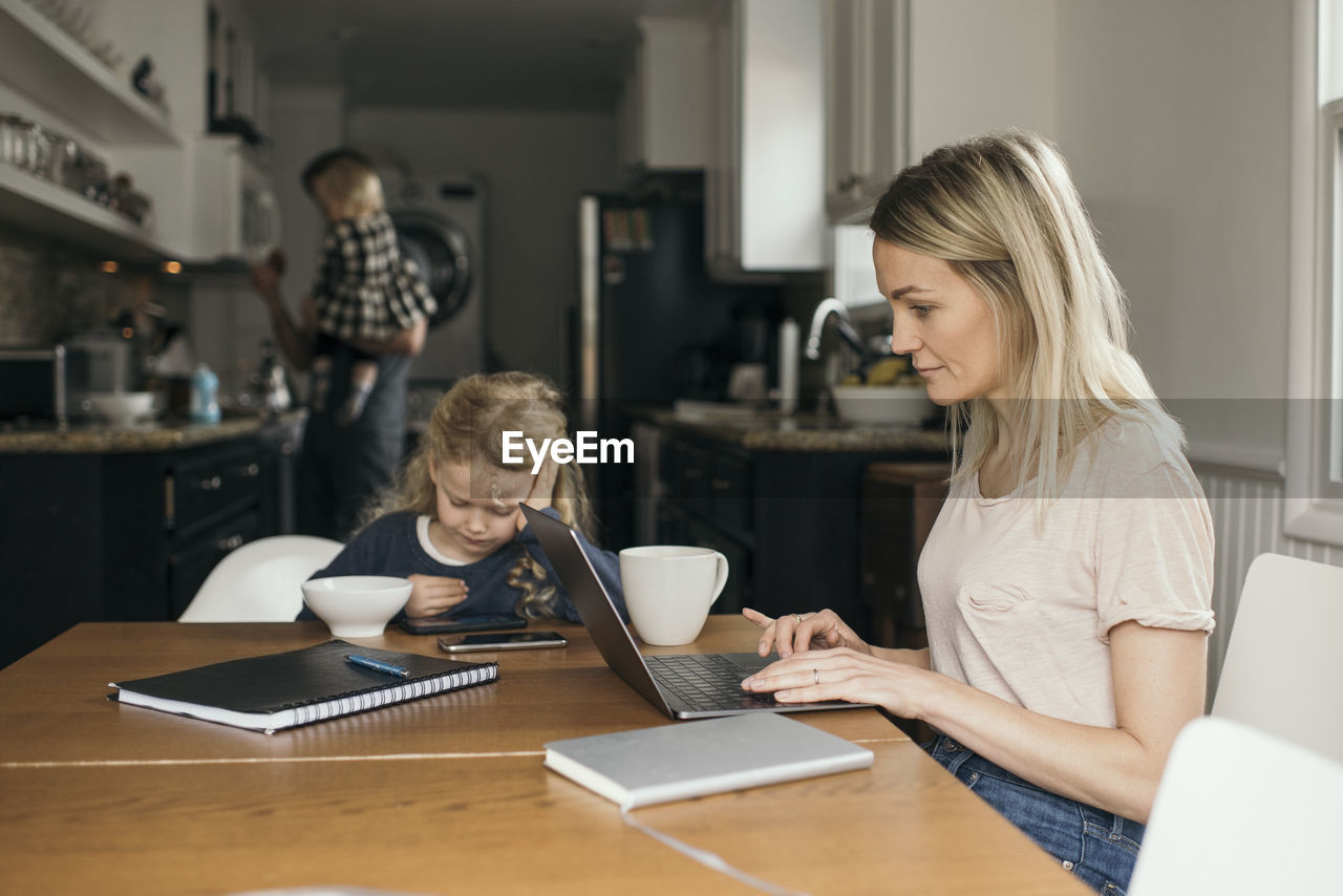 Woman using laptop while daughter having breakfast with family in background at home