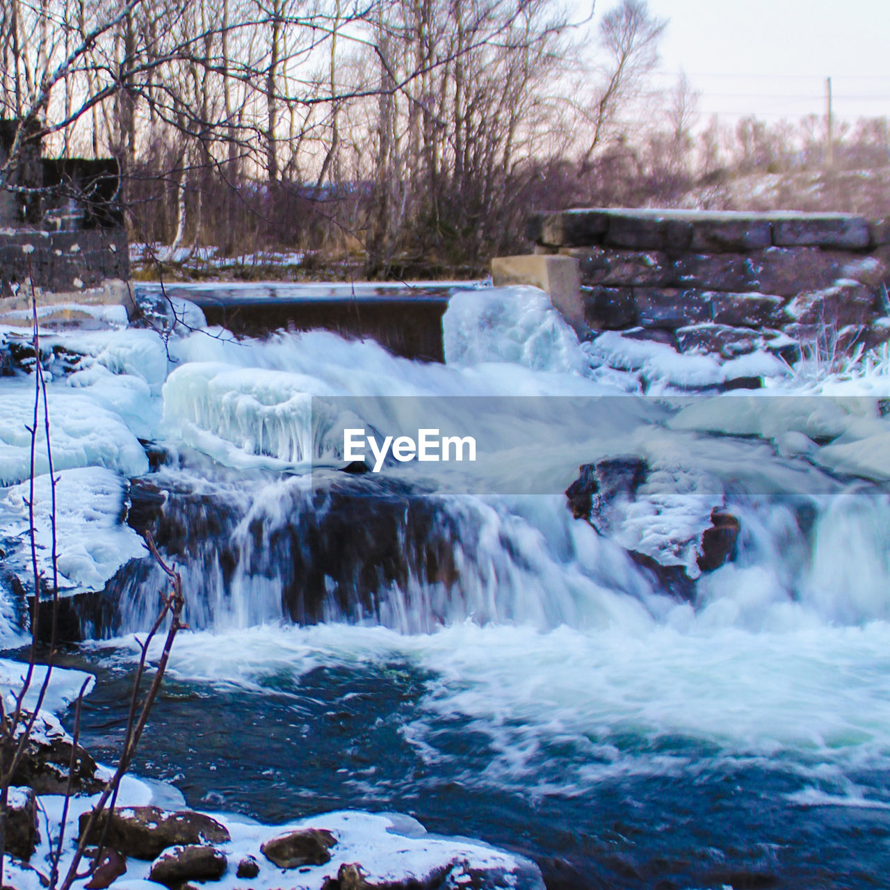 Flowing stream against bare trees against sky during winter